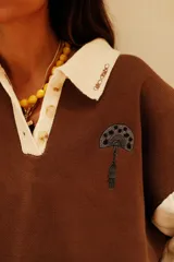 Buzo de punto en color marrón con cuello polo blanco y mangas largas blancas con textura de ochos. Presenta un diseño gráfico de gran tamaño en la espalda en tonos rosado, blanco y negro.