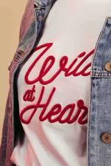 Remera blanca de algodón con cuello redondo y estampado frontal con la frase "Wild at Heart" en letras rojas.