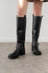 Bota de caña alta color negro, con hebilla y arnés decorativo en el tobillo, y taco bajo. Confeccionada en material sintético PU (poliuretano), libre de componentes de origen animal.