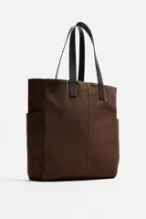 Bolso tipo shopper marrón de tejido de algodón con asas de cuero. Cuenta con dos bolsillos laterales exteriores y un bolsillo interno. Marca Mango.