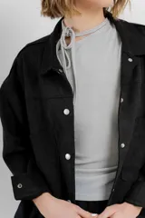 Chaqueta color beige de corte recto, estilo camisa, confeccionada en gamuza con elastano. Presenta cuello camisero, cierre frontal con botones metálicos, bolsillos grandes en el frente y charreteras en los hombros.