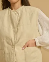 Chaleco corto color beige, con cuello redondo, cierre frontal con un botón y dos bolsillos delanteros.