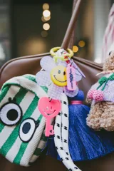 Llavero con aro metálico violeta y dijes colgantes: flor a cuadros lila y blanco con carita sonriente amarilla, cadena de eslabones verde flúor, etiqueta rosa con texto y llave rosa con carita sonriente. Incluye borla de hilos azul eléctrico.