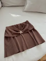 Minifalda marrón texturizada con detalle de estrella de mar dorada en la parte delantera. Presenta una cintura alta con un diseño drapeado.