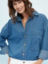 Camisa de denim negro con corte oversize, cuello clásico, cierre frontal con botones metálicos y bolsillo de parche en el pecho.
