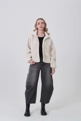 Campera de poliéster tipo sherpa en color natural, con diseño holgado, cuello alto y cierre frontal completo. Presenta un bolsillo de parche con cierre en el pecho y mangas largas con puños elásticos.