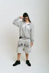 Bermuda de felpa color gris claro con efecto lavado (washed). Presenta un corte ancho con cintura elástica y cordón beige. El diseño frontal incluye un estampado serigrafiado en negro con texto y motivos florales/ornamentales en bordado blanco. Posee remaches metálicos cromados en los laterales y bolsillos.