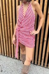 Vestido corto de gasa con estampado de rayas verticales rojas y blancas. Presenta un escote pronunciado en V, diseño halter y drapeado lateral en la cadera con un lazo largo ajustable. Incluye un lazo adicional a juego para usar como accesorio.