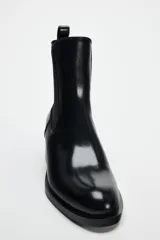 Botín Chelsea de cuero negro con elásticos laterales, puntera redondeada y detalle de placa metálica dorada en el tacón.
