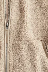 Campera de sherpa suave en color beige, de corte holgado con cuello elevado y cierre frontal bidireccional. Presenta bolsillos frontales tipo canguro y detalles de ribetes en tejido revestido.