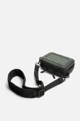 Bandolera rígida negra con formato de caja. Tiene un compartimento principal con cierre de cremallera, un bolsillo interior plano y asa de hombro ajustable y extraíble.