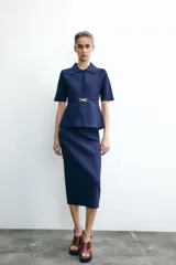 Chaqueta azul marino de punto con cuello solapa y manga corta. Incluye cinturón fino con hebilla dorada y cierre frontal con botones a tono.