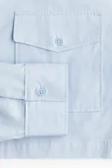 Camisa cropped celeste de algodón con cuello, botones ocultos, canesú con pliegue en la espalda, corte holgado, mangas largas, bolsillos superiores con solapa y botón, puños abotonados y bajo recto.