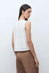 Chaleco sin mangas color blanco crudo, tejido a crochet o punto abierto, con escote en V y cierre frontal mediante lazos cruzados.