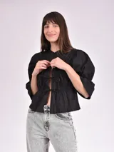 Blusa negra con escote mao y mangas 3/4 con volantes. Presenta detalle de frunces en el frente y lazos para ajustar.