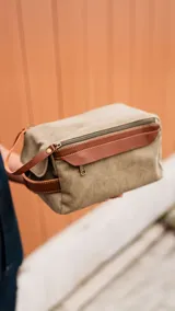 Neceser verde de tela canvas con detalles en cuero marrón. Tiene doble cierre superior, bolsillos internos con cierre y abiertos, bolsillo frontal con solapa y asa lateral.