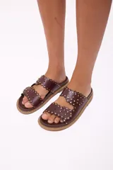 Sandalia tipo mule de mujer, color dorado metalizado, con dos tiras anchas superpuestas con borde festoneado y tachas doradas. La suela es gruesa y de color marrón claro.