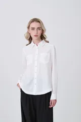 Camisa blanca de manga larga con cuello clásico, dos bolsillos frontales con tapa y botones a presión. Presenta un tejido con textura sutil de rayas verticales.