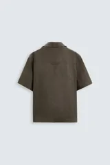 Camisa color marrón oscuro de corte relaxed fit, confeccionada en tejido de lino. Tiene cuello bowling, manga corta, bajo con aberturas laterales y cierre frontal de botonadura.