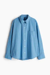 Camisa celeste y blanca a rayas verticales, de corte amplio y mangas largas. Tiene cuello clásico, cierre frontal con botones y bolsillo de parche en el pecho.