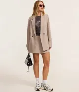 Blazer largo color beige, de corte recto, con cuello solapa, cierre frontal con doble botón, bolsillos tipo ojal y mangas largas con botones decorativos en los puños.