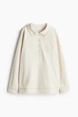 Buzo color crema de algodón con cuello camisero, tapeta de botones y mangas raglán. Tiene un pequeño bordado en el pecho y ribete desflecado en puños y bajo.