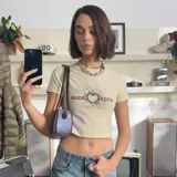 Camiseta corta de manga corta, color beige claro, con cuello redondo y ajuste al cuerpo. Presenta un estampado frontal con un corazón roto que simula un impacto, rodeado de fragmentos, y el texto "NUDE" en rojo a la izquierda y "JECT®" en rojo a la derecha.