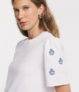 Remera blanca de algodón con cuello redondo, mangas cortas abullonadas y pequeños bordados florales azules en las mangas.