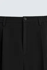 Pantalón negro de corte ancho con cintura elástica, pliegues frontales, bolsillos laterales y traseros de vivo. Tiene detalle de hebilla metálica en la trabilla y cierre frontal con cremallera y botón.