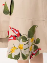 Pollera midi de gabardina color beige con estampado de flores y bordados. Presenta un cruce diagonal con botón dorado personalizado y botón interno para sostener la solapa inferior. Incluye bolsillo plaqué en delantero orientado a cintura y volado en forma de caracol en el ruedo.