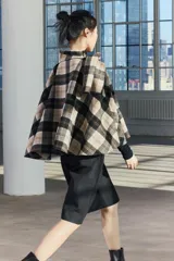Capa de cuello alto confeccionada en tejido con lana a cuadros en tonos beige, negro y marrón. Tiene bajo asimétrico y cierre en la espalda con botón.