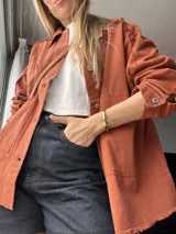 Sobre camisa color ladrillo con cuello camisero, cierre frontal con botones metálicos y bolsillos de parche en el pecho.