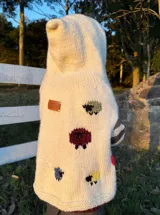 Poncho tejido a mano en lana merino color crudo, con capucha y mangas largas a rayas horizontales en tonos marrón y gris. Presenta apliques bordados de ovejas de colores.