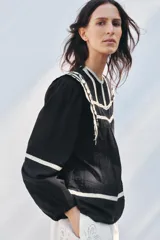 Blusa negra de algodón con cuello redondo y manga larga abullonada. Presenta bordados blancos a contraste en el cuello, hombros y mangas. Cierre en la espalda con abertura y botón.