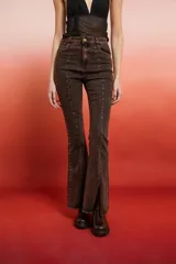 Pantalón de jean de tiro alto, color marrón oscuro con efecto lavado o desgastado, corte flare y tajo en el bajo. Presenta tachas doradas a lo largo de las costuras laterales.