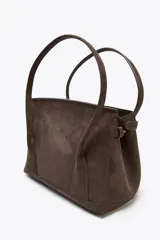 Bolso marrón de gamuza sintética, tipo city bag, con doble asa de hombro y cierre con solapa e imán. Tiene correa ajustable con hebillas metálicas en los laterales y bolsillo interior con cremallera.
