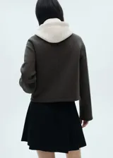 Chaqueta negra corta con capucha gris desmontable y ajustable con cordones. Presenta cuello camisero, cierre de cremallera doble, bolsillos laterales y bajo con goma elástica.