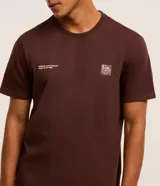 Remera marrón de algodón, corte clásico, con cuello redondo y manga corta. Presenta un pequeño estampado con lettering y un logo en el pecho.