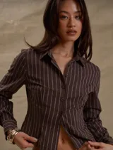 Camisa de manga larga color marrón oscuro con finas rayas verticales blancas. Presenta un corte entallado y ligeramente cropped, con cuello camisero y cierre frontal de botones.