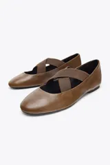 Zapato tipo bailarina plana color marrón, confeccionado en piel. Presenta tiras elásticas cruzadas en la parte delantera y acabado en punta redonda.