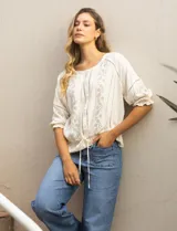 Blusa color crema de viscosa con cuello redondo y abertura central con botones. Tiene mangas 3/4 con puños fruncidos, detalles calados y bordados en mangas, delantera y espalda. La cintura tiene un lazo de ajuste.