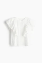 Blusa blanca de algodón con secciones en broderie. Modelo con mangas cortas de volados y cuello redondo con abertura de cerradura y botón oculto en la nuca.