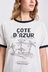 Remera color crudo de manga corta con cuello y puños negros. Estampa frontal con texto "Côte D'Azur" y dibujo de playa con palmeras.