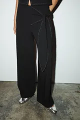 Pantalón negro de tiro medio con cremallera lateral oculta, corte ancho y lazo decorativo en la cadera. Detalle de pespuntes blancos a contraste.