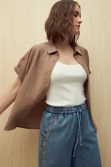 Camisa color marrón claro, de manga corta con puños arremangados, cuello clásico y cierre frontal con botones.