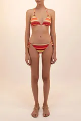 Calzón de bikini a rayas horizontales en tonos naranja, rojo, amarillo y beige. Tiene corte a la cadera, costados sin costuras, argollas doradas y tiras ajustables con detalles metálicos en las puntas.