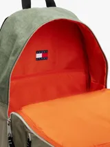 Mochila de nailon color verde oliva, con diseño liso y paneles texturizados. Presenta un compartimento principal con doble cremallera, un bolsillo frontal con cremallera y un parche con el logo de Tommy Jeans en la parte inferior. Las correas y el asa superior son negras.