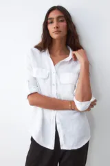 Camisa blanca de lino con cuello clásico, cierre frontal con botones, mangas largas con puños abotonados y dos bolsillos de parche en el pecho.