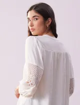 Blusa blanca de gasa con cuello a la base, escote en V y abertura central con botones. Tiene mangas 3/4 amplias con apliques de crochet y detalles plisados en la parte delantera.