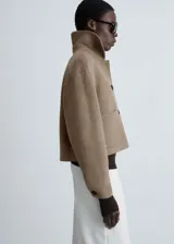 Chaqueta corta color marrón claro, de corte recto y cuello alto. Presenta cierre frontal con botones, dos bolsillos de parche delanteros y puños con botones funcionales.